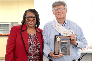 Library District director Shaney Livingston in a red suit stands next to library specialist Sid Jones as Jones holds a plaque honoring his 36 years of service to the Library District during a retirement party for Jones.