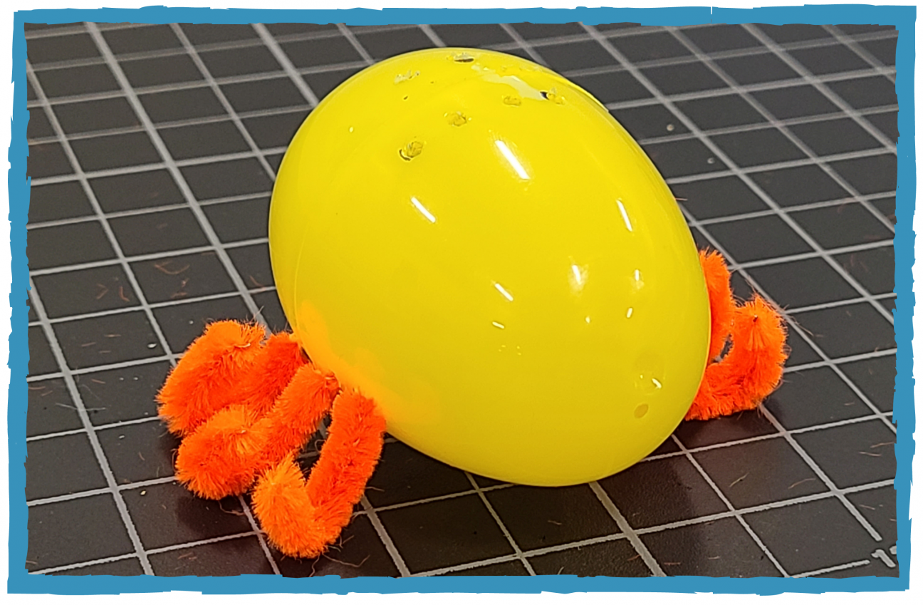 A yellow plastic egg sits on a cutting mat. It has holes cut into the top and side of it, with orange pipe cleaners threaded through the sides. 