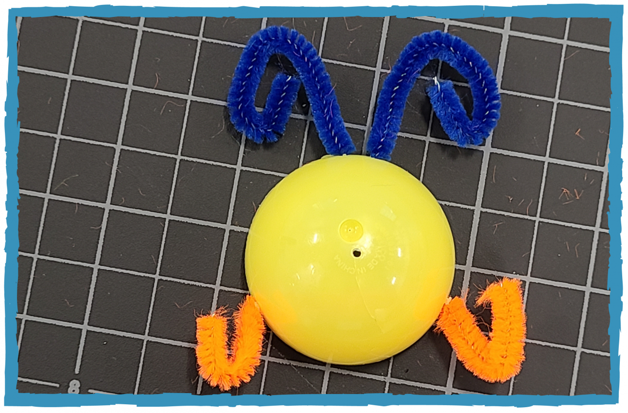 The short half of a yellow plastic egg, with orange pipe cleaners threaded through the sides, and blue pipe cleaner threaded through the top. It looks a little like a bug.