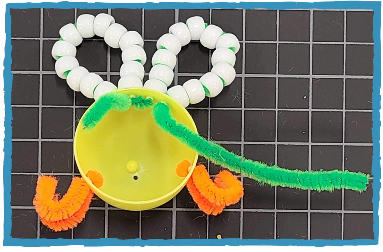 Half of a yellow plastic egg. There are orange pipe cleaners threaded through holes on the sides. Green pipe cleaners are threaded through holes in the top, with pony beads threaded on them, forming the shape of wings.