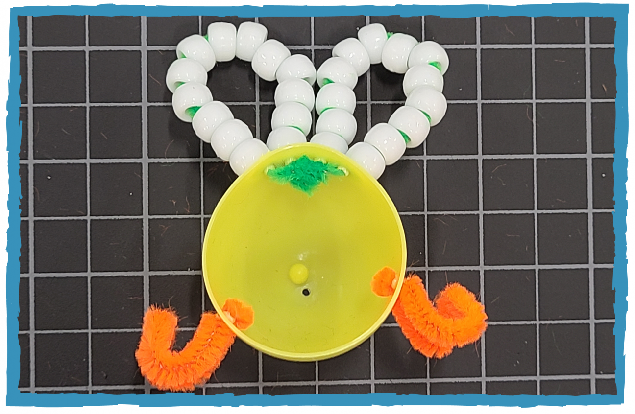 Half of a yellow plastic egg. Orange pipe cleaners are threaded through the sides, with green pipe cleaner threaded through the top. There are white pony beads on the green pipe cleaners, forming wings.