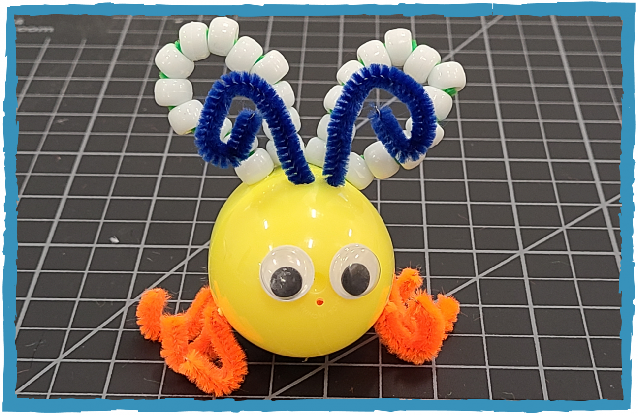 A yellow plastic egg is made to look like a bug. It has six orange pipe cleaner legs, blue pipe cleaner antenna, and wings made of green pipe cleaner and white pony beads.