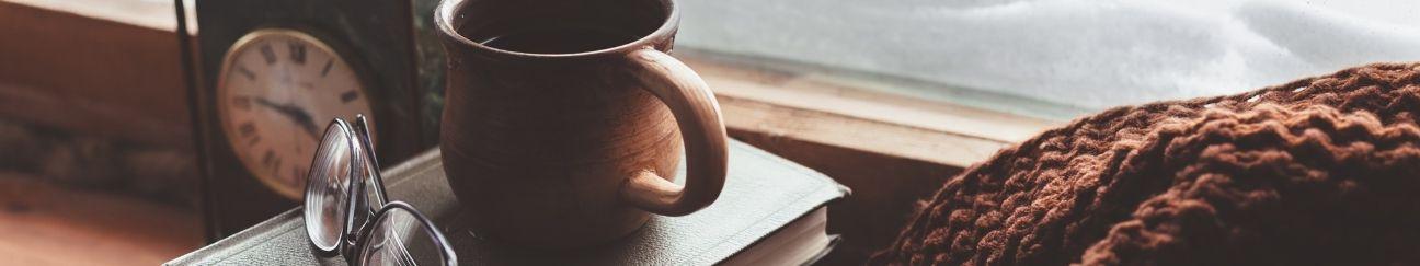 a stack of books with a mug of coffee and a pair of glasses on top with a clock and a sweater next to it facing an open window