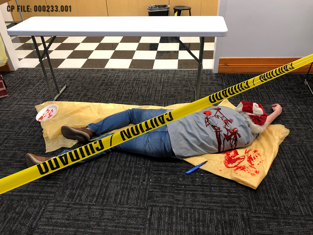 Evidence Photo 001 image of a dead stabbed body lying on a tablecloth. The image is cut through with police tape.