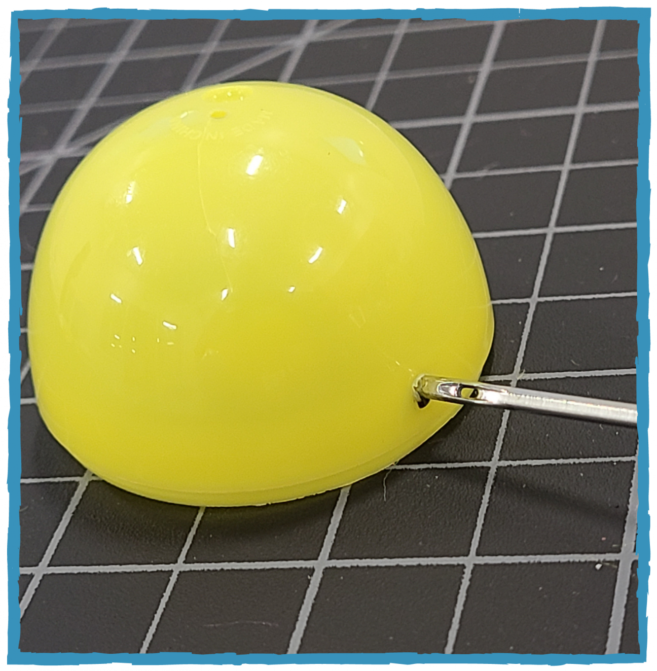 The short half of a yellow plastic egg sits on a cutting mat. There is a needle inserted into a marked hole.