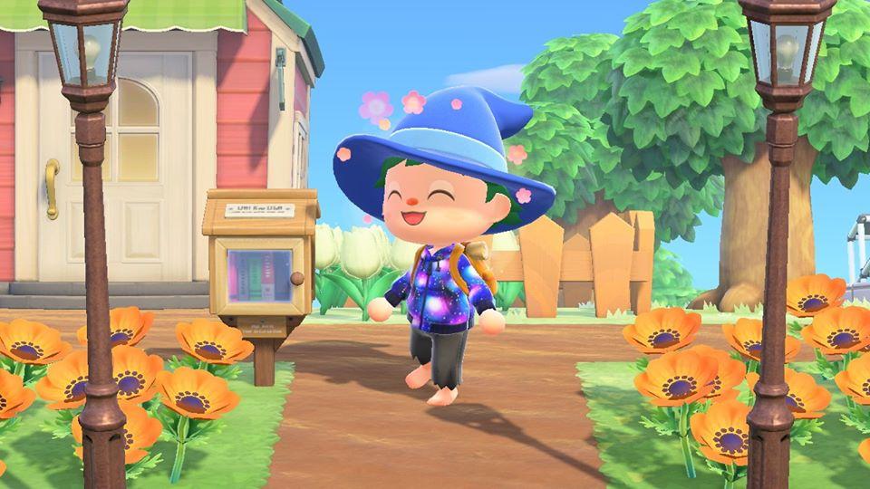 Screenshot of a player character in Animal Crossing: New Horizons. Player character is wearing a witch hat, a galaxy hoodie, and using the Joy Reaction. Player character is standing next to a Little Free Library and flowers.