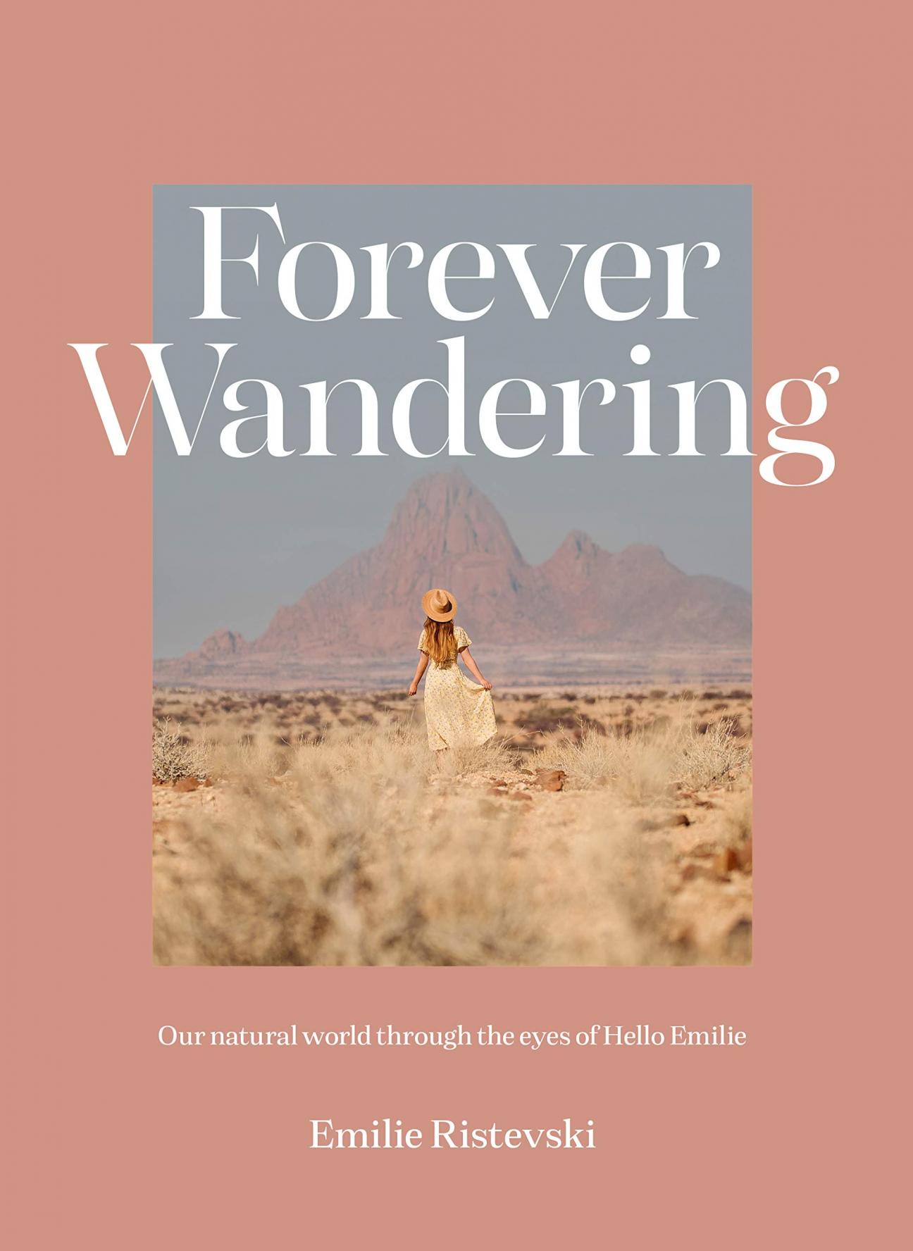 The cover of "Forever Wandering" by Emilie Ristevski, which has a pale pink border surrounding a photograph of a woman in a long dress and straw hat standing in a desert-like area with her back to the camera, looking at a mountain in the far distance.