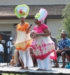 An African American woman dressed in a colorful dress with men playing drums behind her.