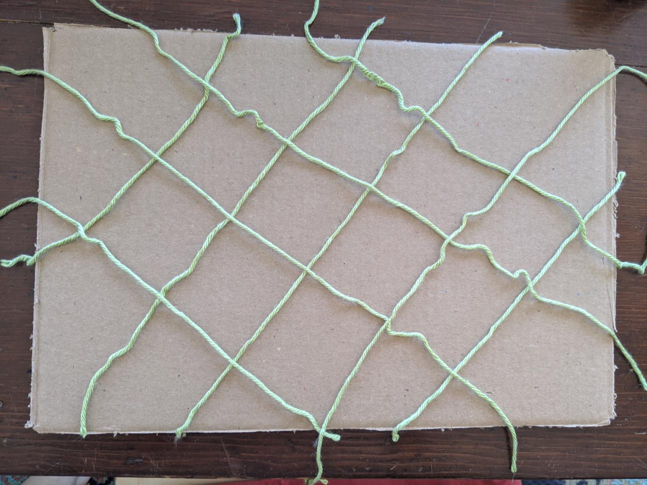 A rectangular piece of cardboard with strands of green yarn criss-crossed over the top of it.