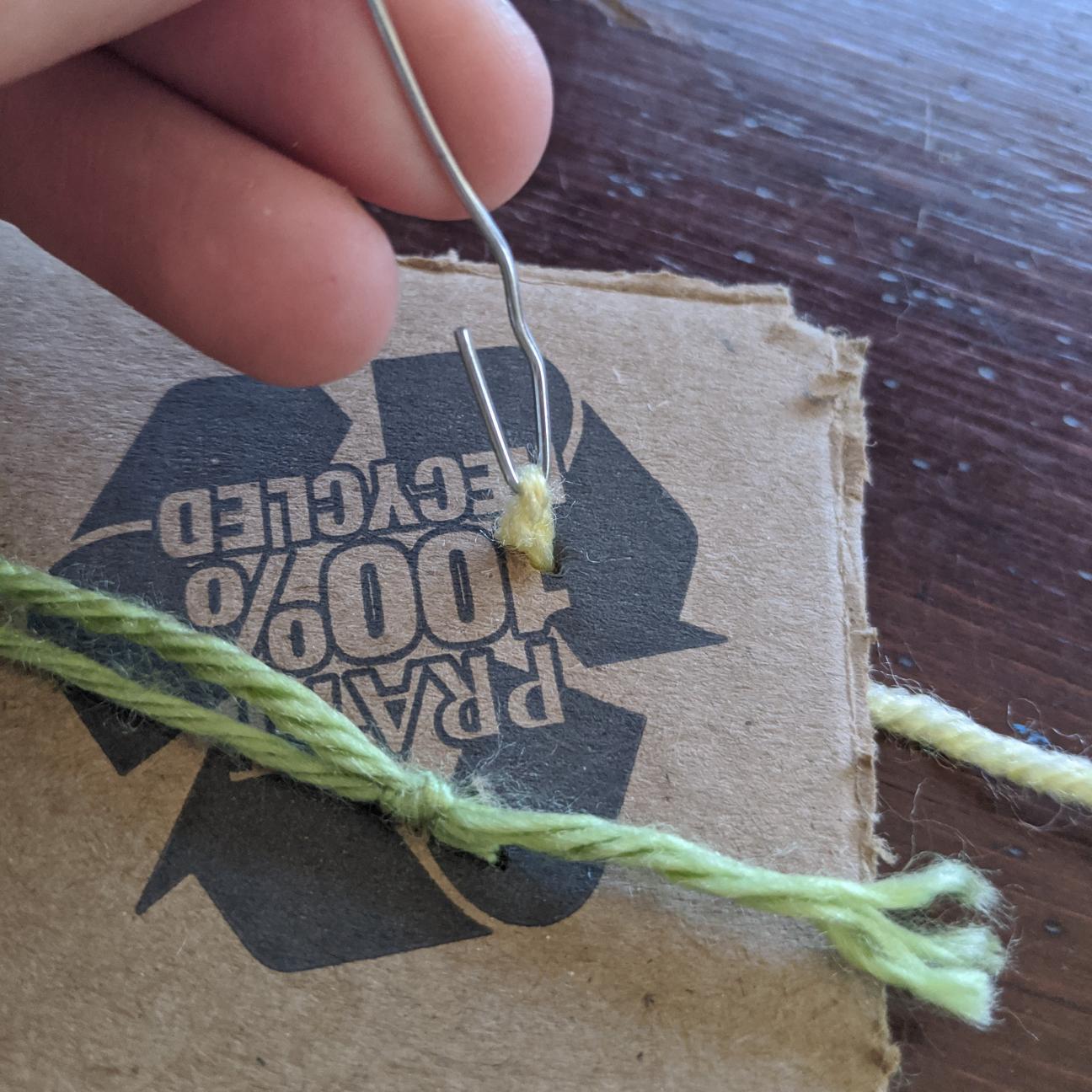 A strand of yarn that has been pulled through to the back of the cardboard using the paperclip hook.