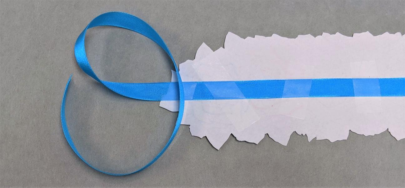Adding ribbon A photo of the backside of a straight flower crown, with blue ribbon taped down along the length of the crown