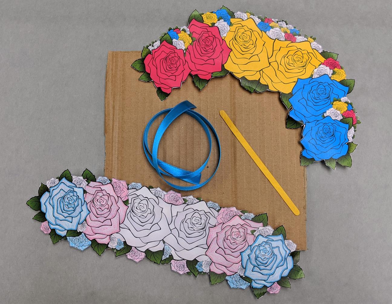 A photo of cardboard, two flower crowns, blue ribbon, and a yellow popsicle stick on a gray background.