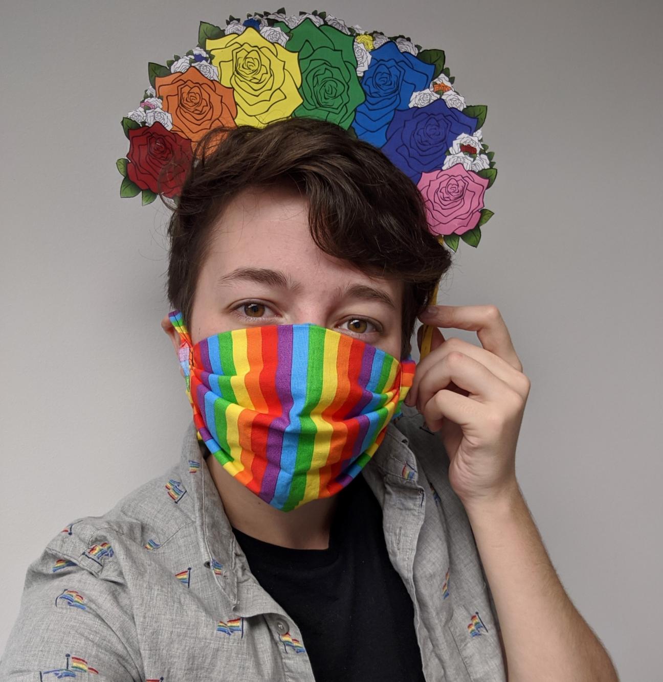 A selfie of a person wearing a rainbow mask and a button down shirt with rainbow flags on it. They are holding a rainbow flower crown above their head.