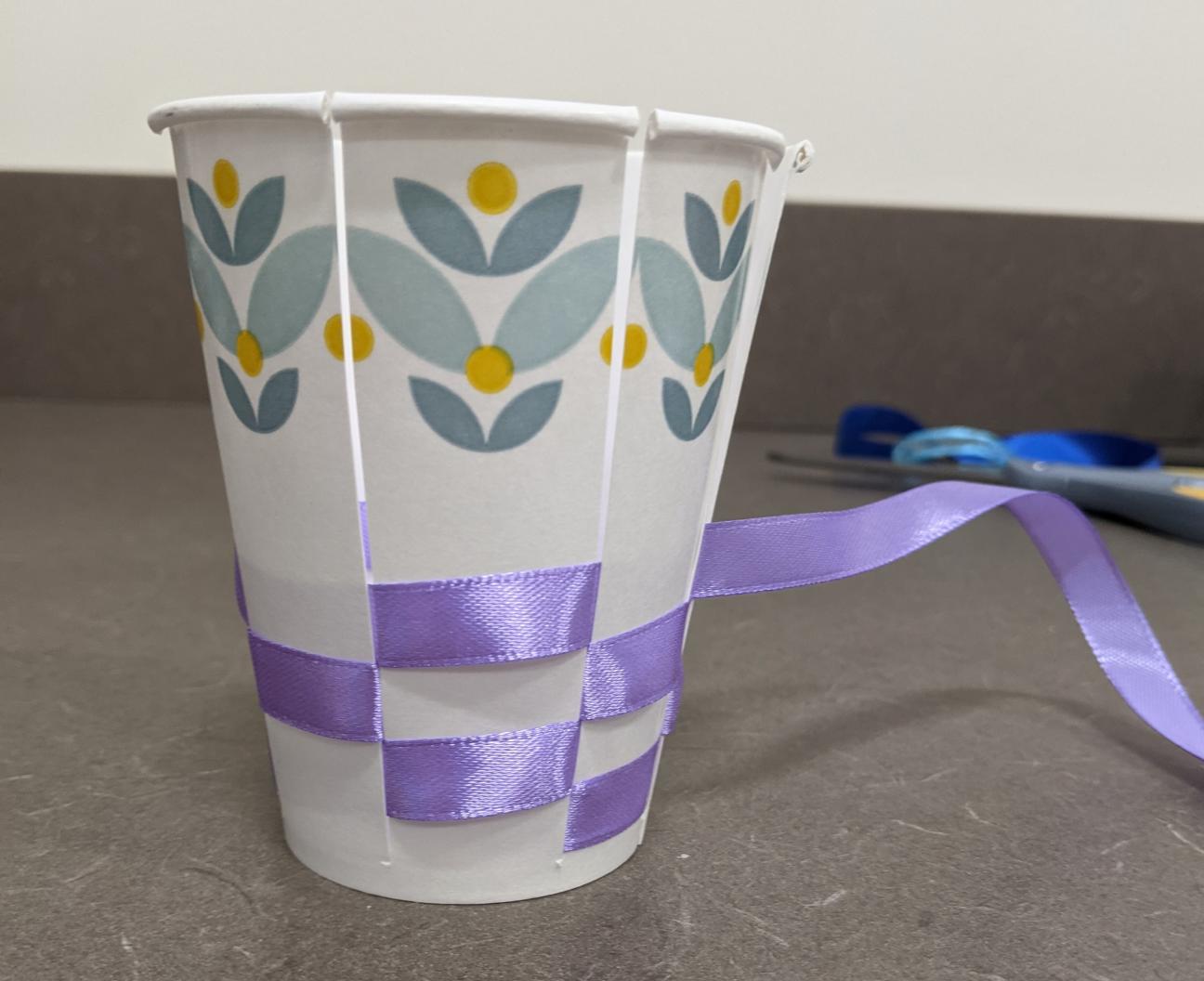 Weave your ribbon Weave in the ribbon. The ribbon pictured is purple and the cup is white with blue and yellow flowers at the top.