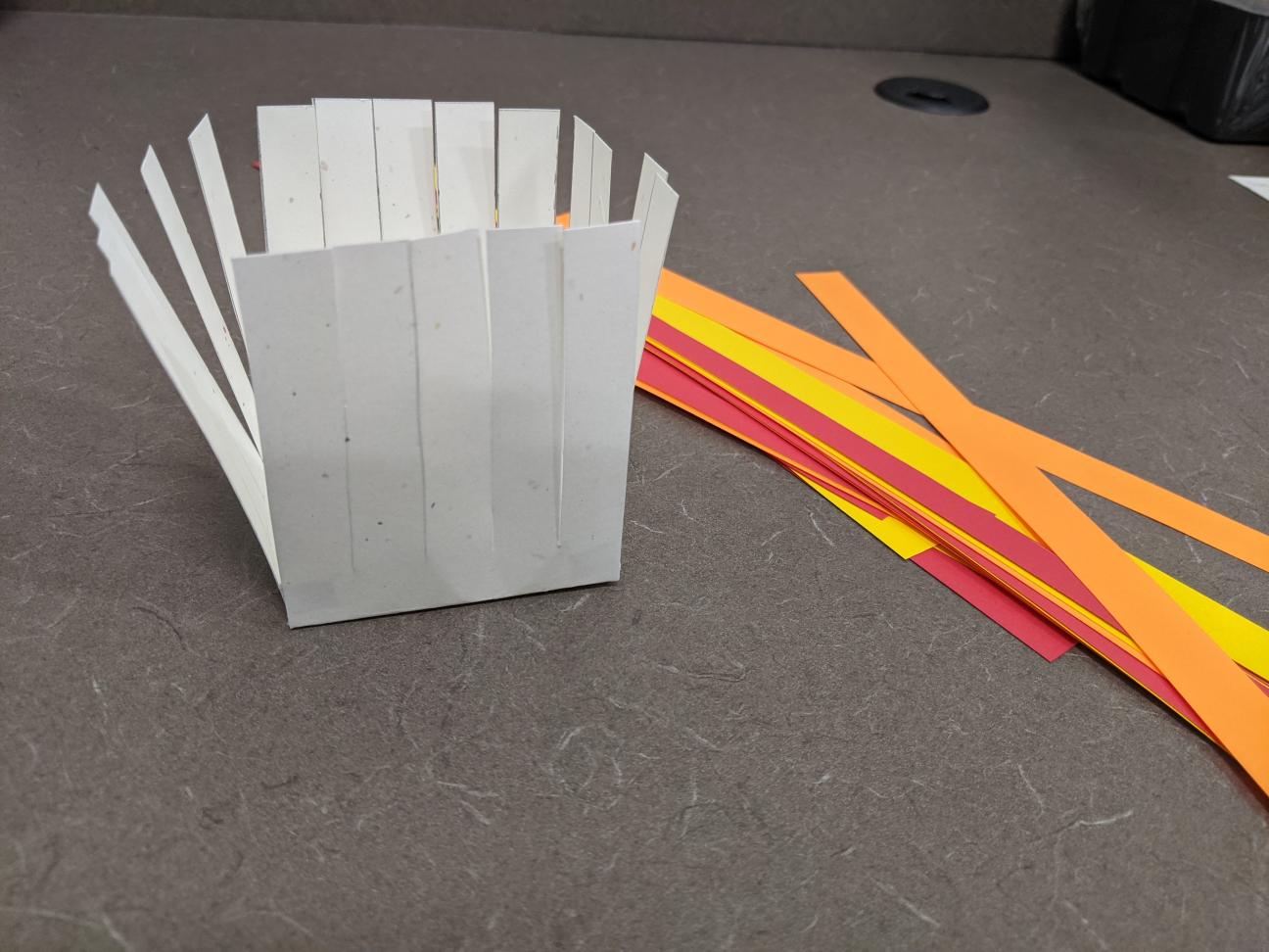 Your basket and paper strips The glued together paper basket, now with thin strips of red, orange, and yellow paper next to it