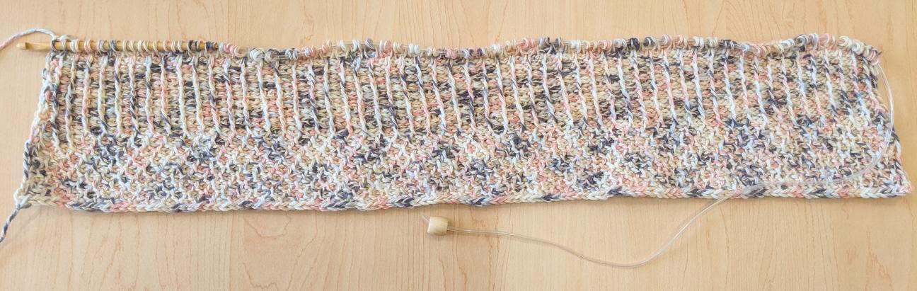 A photo of a baby blanket that is being created. The yarn is pink, blue, and tan, and the hook is at the end of the forward pass. 