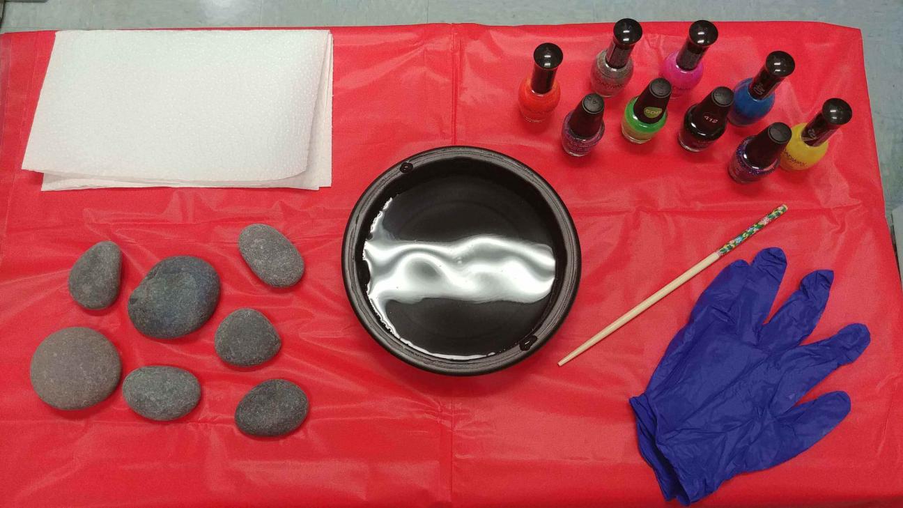 A photo of rocks, a dish of water, nail polish bottles, blue gloves, and a toothpick on a red vinyl tablecloth
