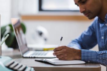 A man writing with a pen in a notebook with a laptop on the table as well.