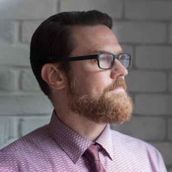 Side profile of white man with a beard, glasses, and red dress shirt and tie looking off into the distance.