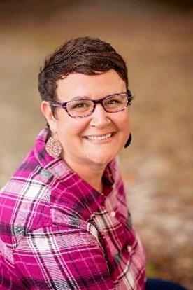 White lady smiling with short dark hair, glasses, earrings, and a pink plaid shirt in front of a brown backdrop.