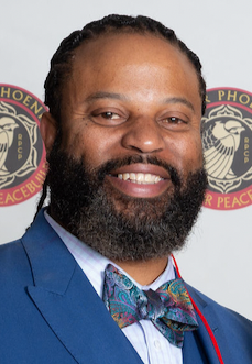 African American man smiling in blue suit with bow tie in front of white background with River Phoenix Center for Peacebuilding logos.