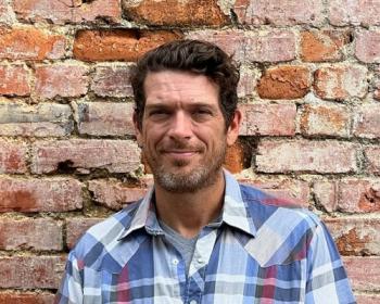 White man with a plaid shirt on smiling and standing in front of brick wall. 