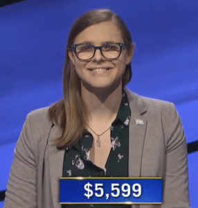 Image of Kate Freeman on the Jeopardy! set -- she wears a trans flag pin on her left lapel. Her total winnings, $5599, are also shown.