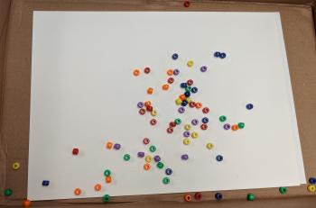 Dropping Dice Method A picture of a piece of paper in the cardboard lid of a paper box. The paper is covered in multicolored pony beads, and light outlines have been drawn around groups of beads.