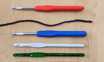 A photo of four crochet hooks and a strand of brown worsted weight yarn. The top crochet hook is an I-9 with a handle, while the bottom three are all K-10.5 hooks with various handle options