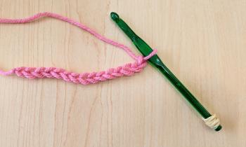 A photo of the green crochet hook with the rubber band at the end, with the beginning of a swatch in pink yarn. It is 15 chains long 