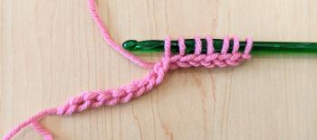 The green crochet hook and pink chain, this time beginning the forward pass. There are seven loops of yarn on the crochet hook.