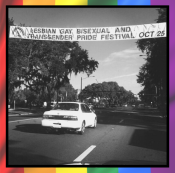 A black and white photograph of a Gainesville street.  There is a white car driving away from the camera, and above it is a banner, strung across the street, stating "Lesbian Gay Bisexual and Transgender Pride Festival Oct 25" with the Pride Center logo on the left.