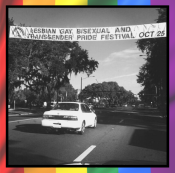 A black and white photograph of a Gainesville street. There is a white car driving away from the camera, and above it is a banner, strung across the street, stating "Lesbian Gay Bisexual and Transgender Pride Festival Oct 25" with the Pride Center logo on the left.